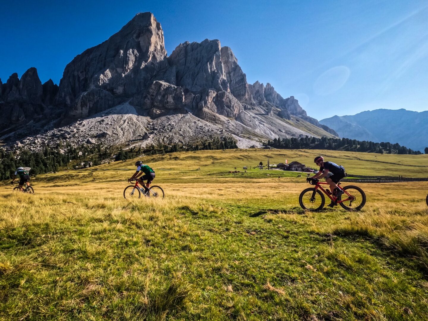 Three cyclists ride gravel bikes across a field in Italy's Dolomites during the YOLOmites5000 bike ride. YOLOmites5000 is a backroads Dolomites cycling route that climbs 5,000 meters in just 80 miles (129 km). Founded by Igor Tavella of Hotel Ustaria Posta, this challenging ride is in September each year and set to the rising and setting of the sun. This is one of the most beautiful road and gravel cycling routes in Alta Badia and indeed in Italy's Dolomites.